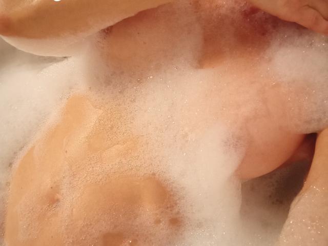 Primo piano del corpo nudo di SharonLeidy immersa in acqua saponata, che mostra il suo seno abbondante e un accenno del suo pene. parzialmente oscurato da bolle bianche. La tonalità della pelle è marrone chiaro..