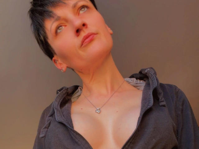 Fotografía de una mujer de pelo corto, atractiva al estilo "NastyMommy", con piel clara, mirando hacia arriba con una ligera sonrisa. Ella lleva una camiseta negra con cremallera, que deja ver parte de su escote y lleva un collar. La luz cálida resalta su rostro..