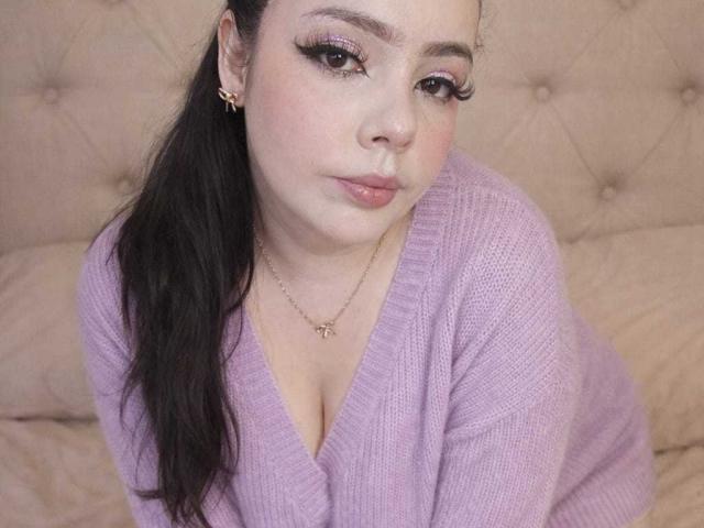 Close-up de NathalyRose69, uma mulher curvilínea com pele clara e cabelo preto em um rabo de cavalo, vestindo um suéter roxo com decote em V. Colar de ouro e brincos. Ela olha de forma sedutora, com um delineador bem marcado..