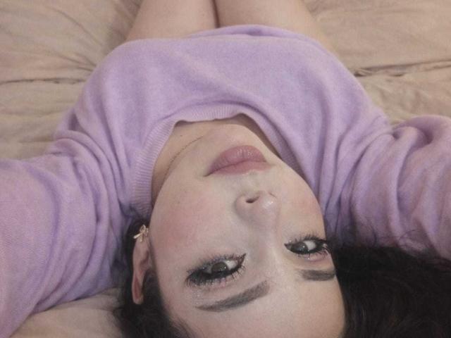 Nahaufnahme von NathalyRose69, die auf einem Bett liegt, den Kopf leicht zurückgelegt, ein lavendelfarbenes Pullover trägt, die Augen halb geschlossen sind, die Lippen leicht geöffnet. teiler Gesicht, dunkle Augenbrauen, dezente Rouge, neutraler Gesichtsausdruck, weiches Licht, intime Pose.