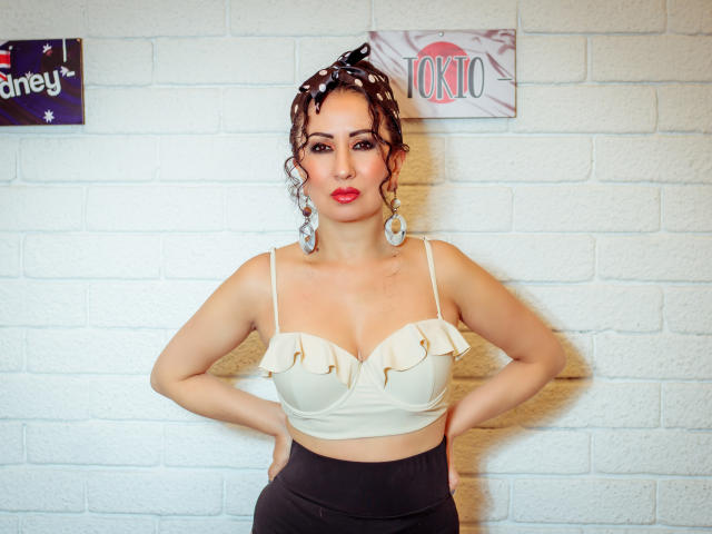 Samantha Antolin se tient devant un mur de briques blanches, les mains sur les hanches, vêtue d'un crop top blanc à volants et de pantalons noirs. pantalon taille haute. Elle a les cheveux bouclés, du rouge à lèvres et de grandes boucles d'oreilles, et elle dégage un charme sexuel confiant..