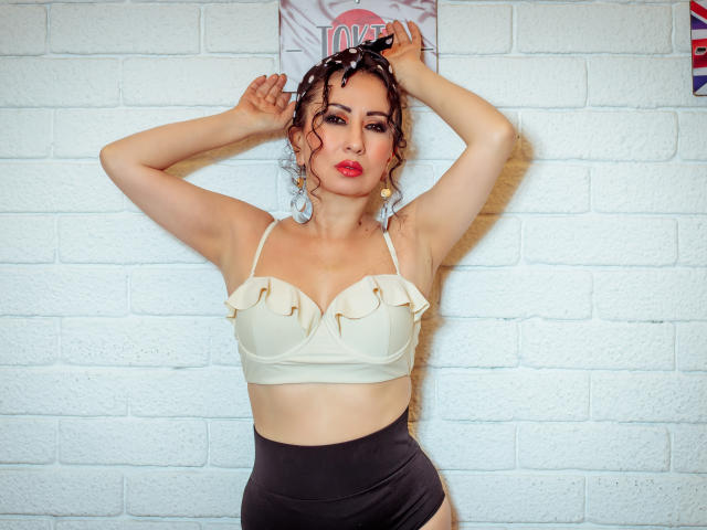 Samantha Antolin, met krullend bruin haar, rode lippenstift en een witte, gerimpelde beha, met armen omhoog tegen een witte bakstenen muur. Zwarte shorts met hoge taille, sexy, zelfverzekerde pose, opvallende kleuren..