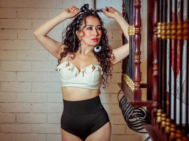 SamanthaAntolin stands in a provocative pose, arms raised, wearing a white bralette with gold studs and black high-waisted panties. She has curly dark hair, red lipstick, and a confident expression.