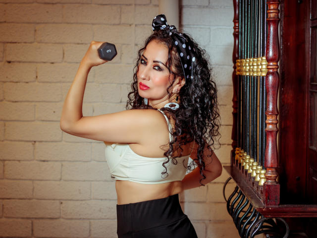 Samantha Antolin, uma latina curvilínea com longos cabelos cacheados pretos, um top esportivo branco, calças pretas de cintura alta, segurando algo cinza. haltere, olhando para trás, lábios vermelhos, laço de cabelo preto, parede de tijolos com pouca luz, piano à direita.