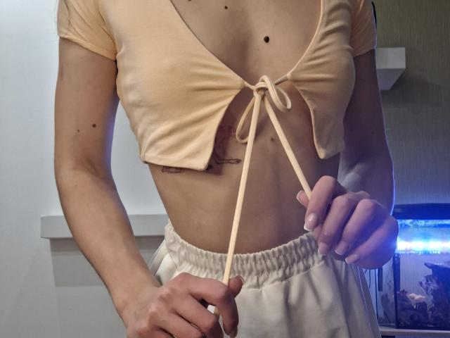 Photograph of a slender woman, MilaYanis, tugging on the tie of her peach-colored, crop top, exposing her midriff. She's wearing white, high-waisted pants. Her hands grip the tie suggestively.