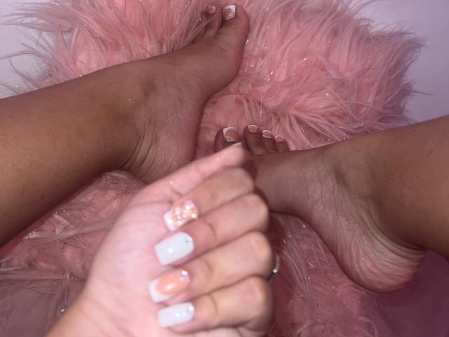 Primer plano de los pies de ChelseaEvans, de piel marrón oscuro, cruzados y entrelazados. Uñas pintadas de blanco, superficie suave y rosada debajo. Dedos ligeramente apretados, creando una atmósfera sensual e íntima..