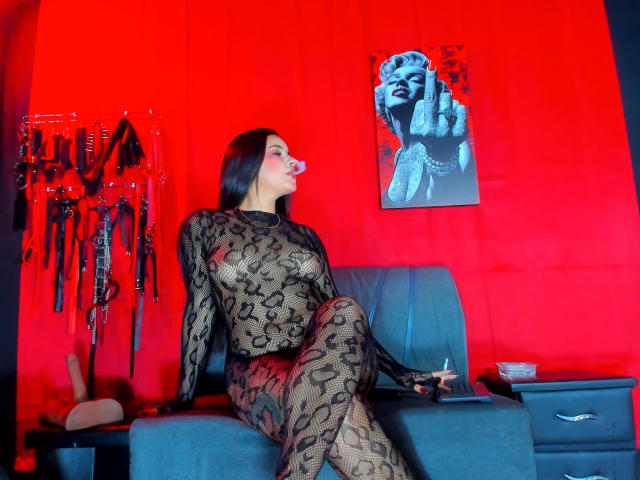 ArtemisaStone, vestita con un abito nero aderente in pizzo, è seduta su un divano nero, di fronte a una parete di colore rosso acceso. Un dipinto erotico, dai colori blu e bianco, è appeso sopra. L'illuminazione rossa esalta la scena..