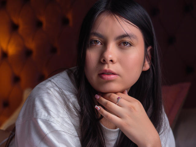 Photo en gros plan de CamiiVasquez, une femme corpulente avec de longs cheveux noirs, portant une chemise blanche et reposant son menton sur sa main. avec une bague à son doigt. Fond sombre et touffu. Expression sérieuse, voire grave..
