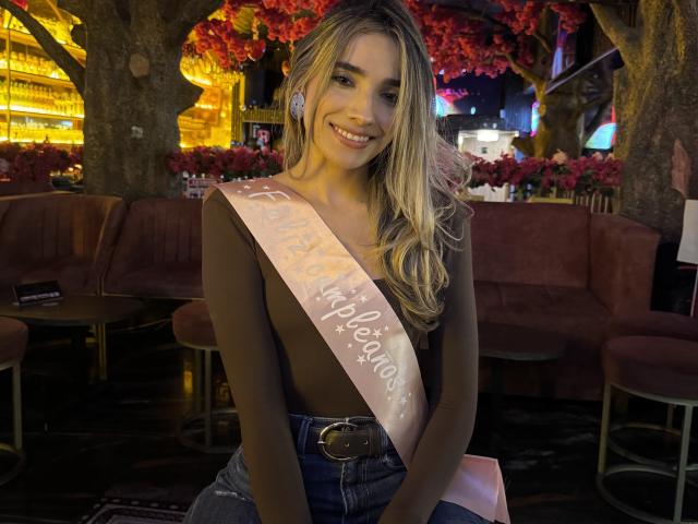 AdharaCooperr sits smiling with long blonde hair, wearing a black top and denim skirt, a pink sash with white text draped over her shoulder. Dimly lit outdoor café at night.