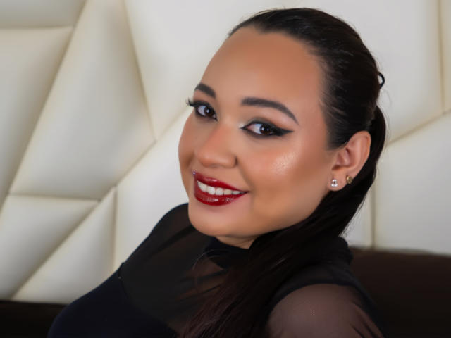 Fotografie von Antonela Gil mit dunklem, zurückgebundenem Haar, die ein durchsichtiges schwarzes Oberteil, roten Lippenstift und Diamant-Ohrringe trägt. mit einem Lächeln in die Kamera blickend, mit strahlenden Augen und glatter Haut. Hintergrund: Weißes, gestepptes Muster.