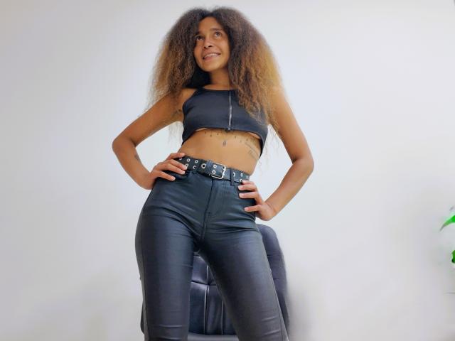 Fotografía de una joven, SweetSexx, con cabello castaño rizado, que lleva un top corto negro y pantalones de cuero negro de cintura alta. pantalones, manos en las caderas, postura segura, fondo blanco brillante, sexy.
