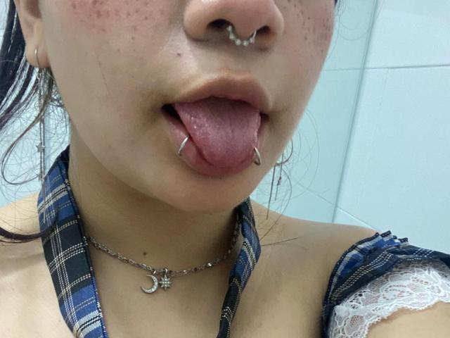 Gros plan d'AmaraKazumi avec un nez et une langue percés, tirant la langue avec un autre piercing. Elle porte un haut à bretelles à carreaux avec de la dentelle et un collier en forme de croissant de lune en argent. Incroyablement sexy..