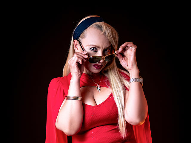 Anastasia Taylor, bionda con i capelli lunghi, indossa un vestito rosso e una fascia nera, e tiene gli occhiali da sole davanti al viso. sfondo nero, luce intensa, scollatura visibile, espressione seducente, alto contrasto.