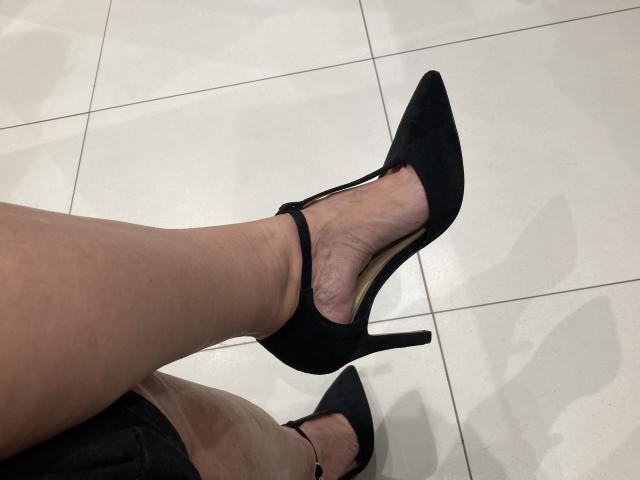 Close-up van de benen van DeesseBleue, die zwarte stilettohakken met enkelbandjes draagt, gekruist bij de enkels, op een witte betegelde vloer. vloer.

de huid heeft een lichte tint en vertoont enige textuur. De hakken zijn puntig en elegant..