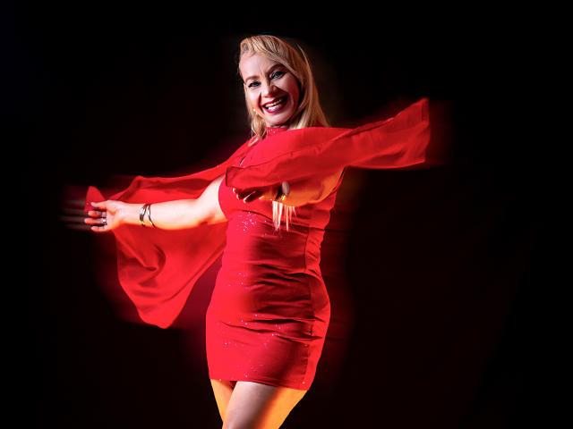 Anastasia Taylor, vêtue d'une robe rouge moulante à manches larges, se tient devant un fond noir. Elle sourit, les bras tendus, et rayonne sous une lumière rouge intense. Ses cheveux blonds sont lisses. Elle est incroyablement séduisante..