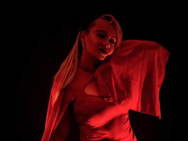 Anastasia Taylor está em pé sob uma iluminação vermelha fraca, vestindo um vestido vermelho transparente que revela sua região do colo. Ela tem cabelos loiros longos e uma expressão sedutora, com o braço levantado. A luz vermelha cria uma atmosfera sensual e sedutora..