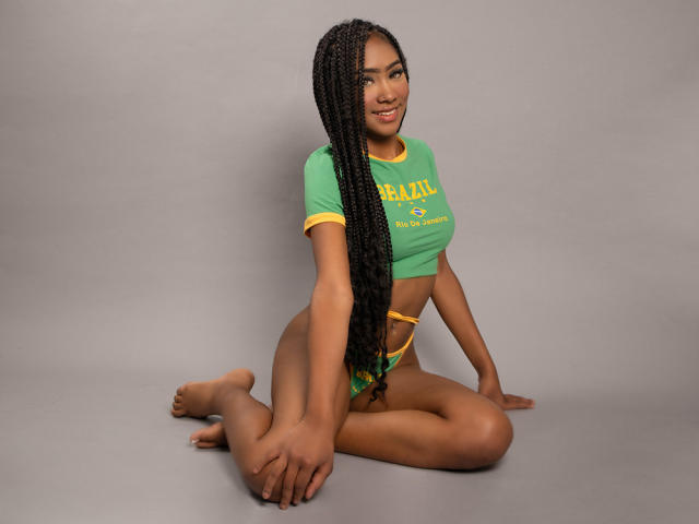 LunaRosee, met donkere huid en lang gevlochten haar, zit op de vloer, benen gespreid, en draagt een groene crop top en een gele onderbroeken, die een getraind lichaam en een zelfverzekerde, sexy glimlach laten zien..