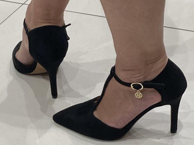 Primo piano dei piedi di DeesseBleue, che indossa scarpe nere con tacco alto e cinturini alla caviglia, decorate con ciondoli dorati. Le scarpe hanno la punta affusolata e tacchi a spillo sottili. La sua pelle è chiara, e si trova su piastrelle bianche..