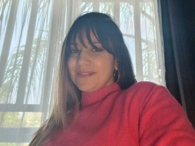 Fotografia de Roxanne Blair, vestindo um suéter vermelho de gola alta, com cabelos castanhos longos e franja, em frente a janela com cortinas translúcidas. Sua expressão é ligeiramente sedutora, com um toque de sorriso..