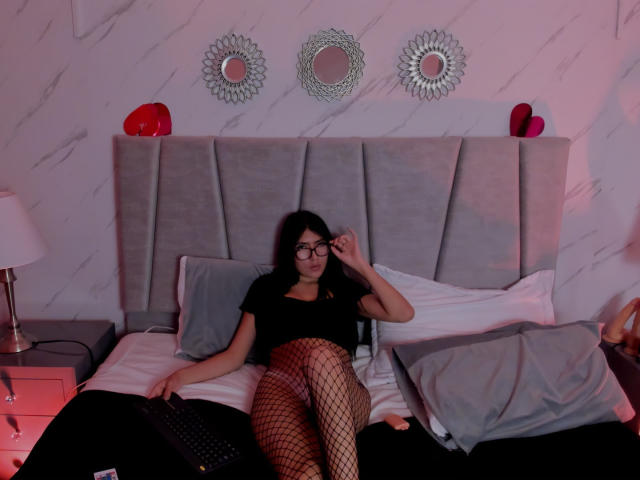 EmmaRouxx sits on a grey, tufted headboard bed wearing black top and fishnet stockings, legs spread, hand on face, red hearts on wall, mirrored decor above. Bedroom setting with red lighting.