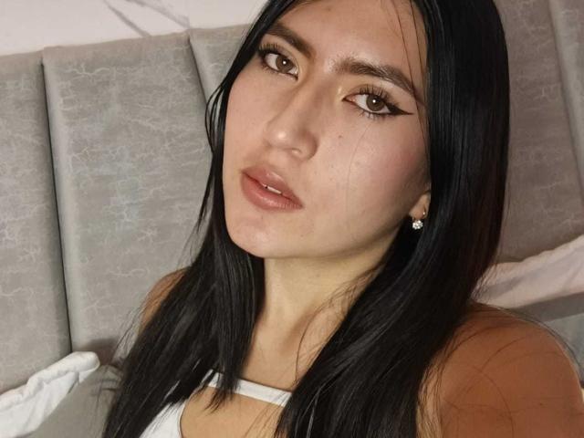 Close-up of EmmaRouxx with long black hair, full lips, and brown eyes, wearing a white top, lying on a gray textured bed. Her expression is sultry, with a hint of sexual tension.