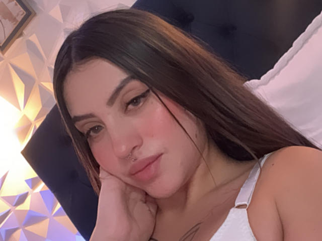 Close-up of LunaaBlossom, a young woman with long black hair, resting her head on her hand. She has a nose ring, thick eyebrows, and a white tank top. Soft lighting, subtle sexual tension.