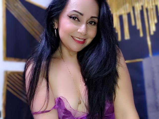 Close-up of Zaria69, a sexy Asian woman with long black hair, red lipstick, and a purple strapless top, smiling seductively. Her chest is partially exposed, showing cleavage. Golden fringe in background.