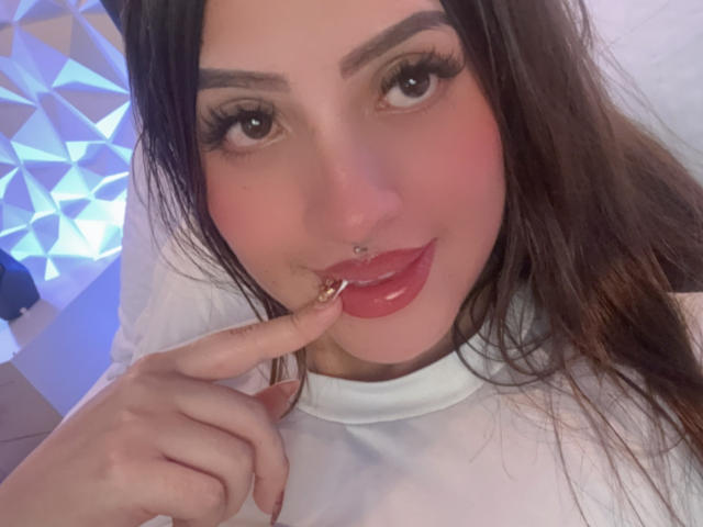 Gros plan de LunaaBlossom, une femme à la peau claire, avec des cheveux bruns foncés, un maquillage prononcé et un piercing au septum. Elle suce son doigt avec ses lèvres rouges et brillantes, et elle porte une chemise blanche. Extrêmement séduisant..