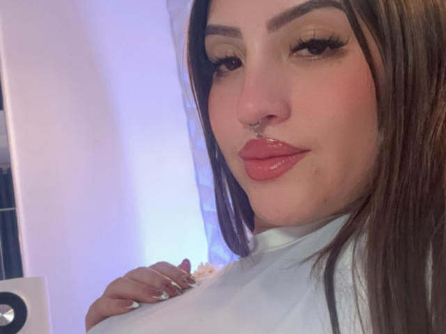 Selfie de perto de LunaaBlossom, uma mulher morena com sobrancelhas grossas, cílios longos, lábios vermelhos e um piercing no nariz. Ela está vestindo uma blusa branca, olhando diretamente para a câmera com uma expressão sedutora..