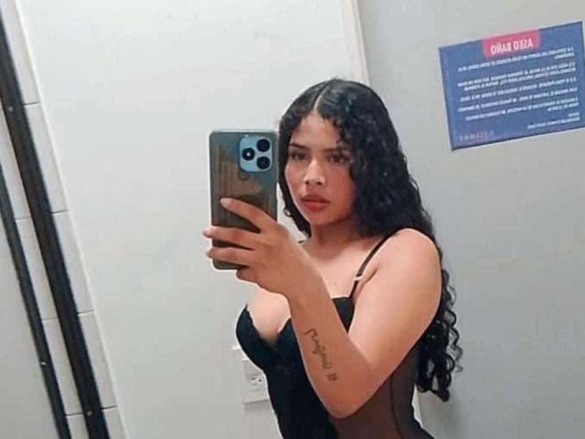 Selfie van AshlyCorines met lang, krullend zwart haar, die een zwarte top met spaghettibandjes draagt en een telefoon vasthoudt voor een spiegel, waarop een tatoeage op haar linkeronderarm te zien is, met een verleidelijke blik..