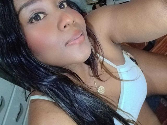 Selfie de primer plano de Yirily, una atractiva mujer asiática con cabello largo y negro, que lleva un top blanco, mostrando su escote y... Collar con girasol. Está inclinada hacia adelante, mirando directamente a la cámara con una sonrisa sutil..