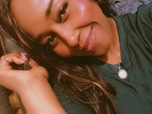 Close-up de Yirily, sorrindo de forma sedutora, com pele brilhante e cabelo castanho escuro. Ela está vestindo uma camisa verde e um colar de girassol, e sua mão está tocando sua cabeça. Muito atraente..