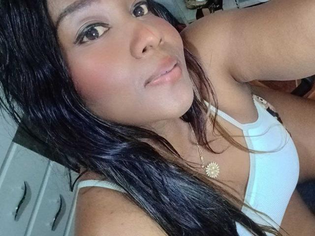 Photo en gros plan de Yirily, avec de longs cheveux noirs, des yeux marrons et une peau brun clair. Elle porte un débardeur blanc, qui dévoile légèrement sa poitrine. Sourire doux, collier avec un motif de soleil. Incroyablement sexy..