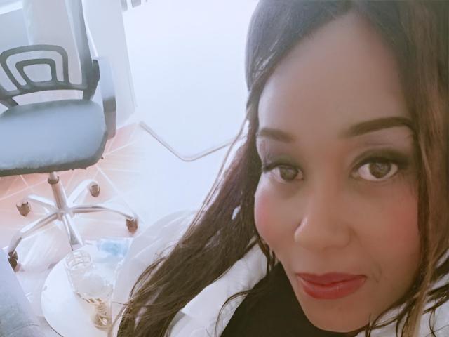 Close-up selfie van Yirily, een Aziatische vrouw met lang bruin haar, rode lippenstift en veel eyeliner, die naar voren leunt. camera, geplaatst op een bureaustoel, achtergrond van een lichte kamer. Heel aantrekkelijk.