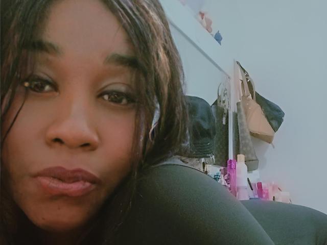 Fotografía de primer plano de Yirily, una mujer de piel oscura con cabello largo y negro, labios carnosos y ojos intensos. Ella está inclinada hacia adelante, ocultando parcialmente un fondo desordenado con luces rosas y ropa colgada..