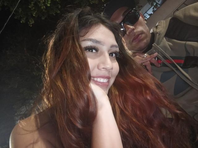 Foto ravvicinata di AnyelinAndMark con lunghi capelli castani ondulati, che sorride mostrando i denti bianchi, con una mano vicino alla bocca. Uomo con barba e occhiali da sole, giacca beige, parzialmente visibile sullo sfondo. Tensione sessuale.