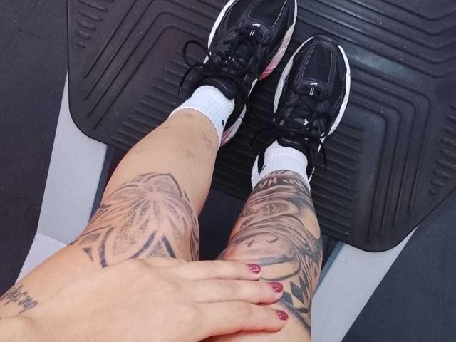 Tattooed legs of AnalisseJade with red nail polish, wearing black sneakers and white socks, resting on a black textured surface. Hand gripping thigh, visible tattoos of flowers and text.