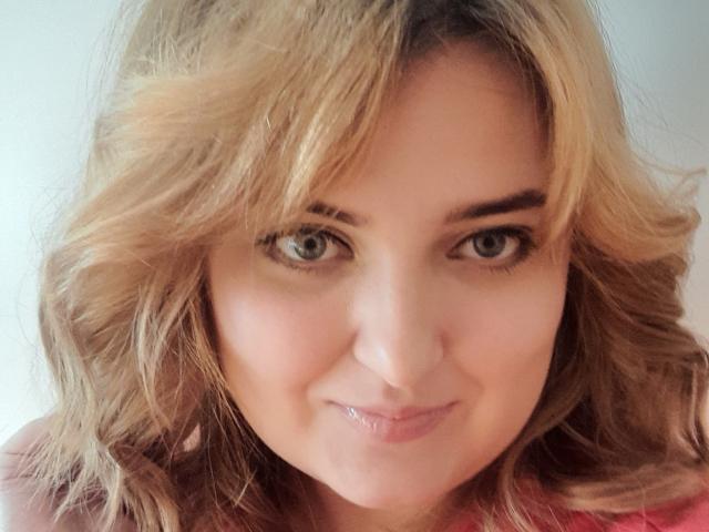 Close-up photo of JuliaNorry, a young woman with light brown, wavy hair and green eyes, smiling subtly. She has a fair complexion, wearing a pink top, with soft natural light. Fucking cute.