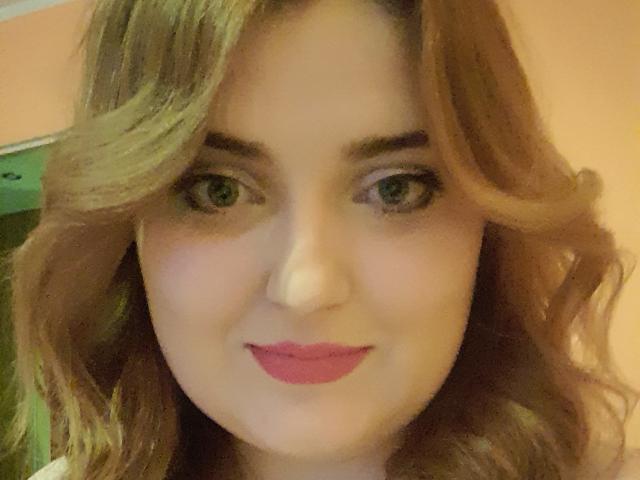 Close-up of JuliaNorry with light brown wavy hair, green eyes, and pink lipstick. She's smiling slightly, with a subtle hint of sexual tension from her direct gaze and bold lip color.