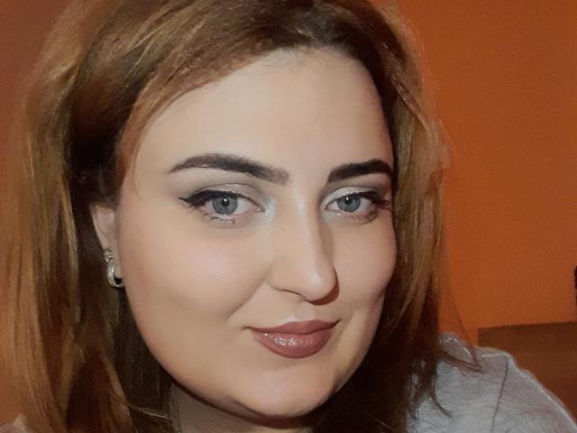 Close-up of JuliaNorry, a young woman with fair skin, blue eyes, and brown hair. She's smiling slightly with glossy pink lips. Her eyelids are lined with black makeup. She's wearing a gray top.