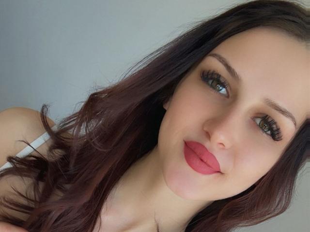 Nahaufnahme von ZoeCarterX, einer weißen Frau mit langen, welligen, dunkelbraunen Haaren, dicken, schwarzen Wimpern und leuchtend rotem Lippenstift. Sie lächelt leicht und schaut direkt in die Kamera. Verdammt heiß..