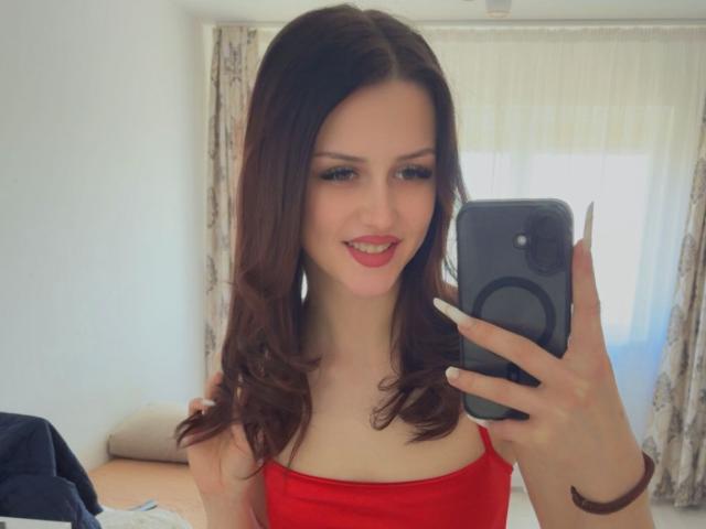 ZoeCarterX prend un selfie devant un miroir, tenant un téléphone noir, et porte un haut rouge sans bretelles, souriant avec des lèvres roses. Longs cheveux bruns, murs blancs, rideaux fleuris. Tellement mignon, d'une beauté sexy..
