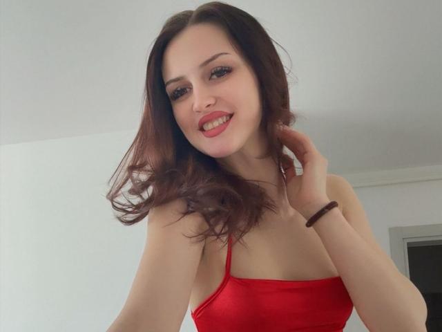 Photograph of ZoeCarterX with pale skin, dark brown wavy hair, red strapless top, smiling with red lipstick, hand touching her hair, visible nipple outline, sexy and confident pose.