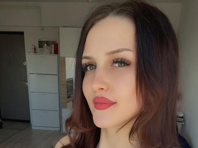 Close-up van ZoeCarterX, een vrouw met bleke huid, lang donkerbruin haar, rode lippenstift en dikke zwarte eyeliner. De achtergrond toont een witte koelkast en een grijze deur. Verdomd aantrekkelijk, met een lichte grijns..