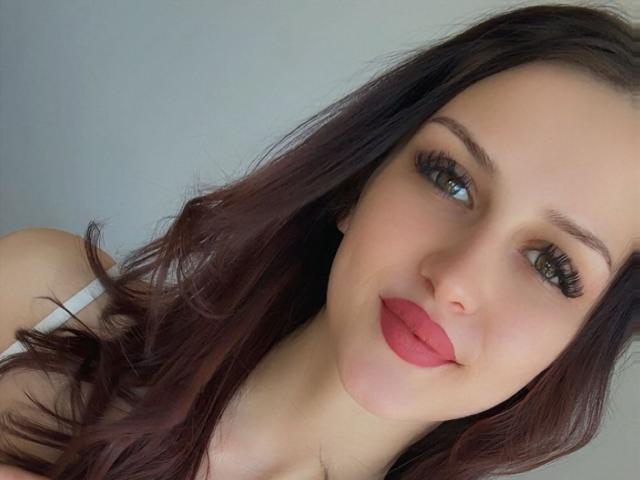 Nahaufnahme von ZoeCarterX, einer hellhäutigen Frau mit langen, dunkelbraunen Haaren, leuchtend rotem Lippenstift und dicken, schwarzen Wimpern. Sie lächelt leicht und blickt mit ihren blauen Augen direkt in die Kamera. Ihre Schultern sind sichtbar..