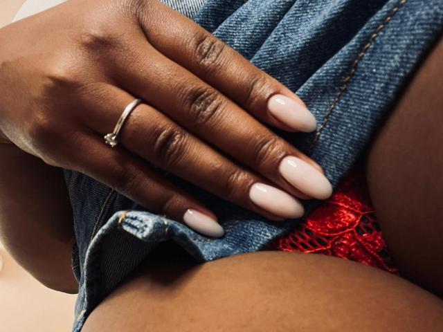 Close-up da mão marrom escura de LisaMendez, com unhas longas pintadas de branco, descansando em sua calça jeans, revelando renda vermelha. calcinhas. Ela usa um anel de prata. Tensão sexual causada pela proximidade física..