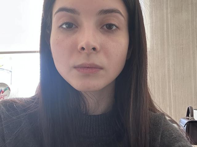 Close-up de Christene Taglauer, uma jovem com pele clara, cabelo castanho escuro e seios pequenos e naturais. Ela está vestindo um suéter preto, olhando diretamente para a câmera com uma expressão neutra. Fundo claro.