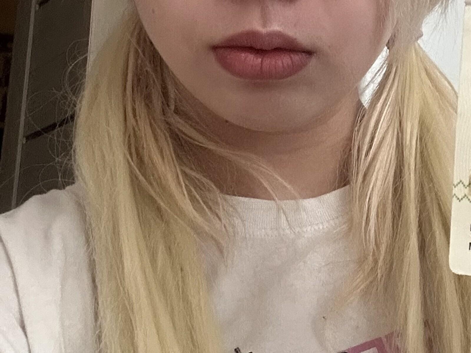 Nahaufnahme von JeanaLamontes Gesicht, die ihre vollen Lippen mit dunkelrotem Lippenstift zeigt. Sie hat blondes, leicht zerzaustes Haar und trägt ein weißes Hemd. Die Beleuchtung betont ihre Gesichtszüge und Lippen..