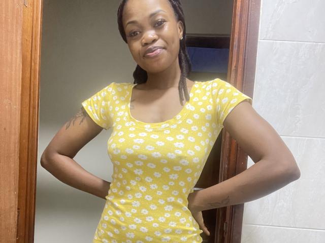 Sashie, une femme noire attrayante, à la peau foncée et aux cheveux tressés, se tient avec assurance dans un encadrement de porte, les mains sur les hanches. Elle portait une robe jaune moulante avec des motifs floraux blancs. Elle sourit légèrement.