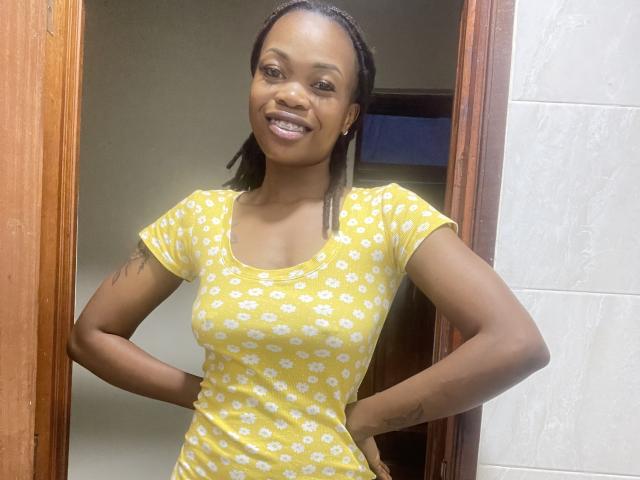 Sashie, an attractive black woman with dark skin and braided hair, stands confidently in a doorway. She wears a tight yellow dress with white flower patterns, smiling broadly. Her hands rest on her hips.